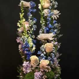 Tall pastel arrangement of peach roses and blue delphinium in a white bowl vase on draped fabric