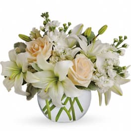 Low arrangement of white lilies, ivory roses, and white stock in a round clear glass vase.