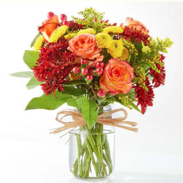 Compact bouquet of orange roses, red mums, and yellow blooms in a clear glass jar with raffia bow.