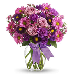 Mixed bouquet of purple and pink flowers with roses in a clear vase tied with a purple ribbon