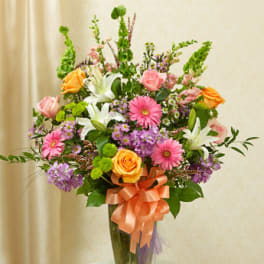 Tall mixed bouquet of pink, orange, and purple flowers with white lilies in a clear vase with a peach bow
