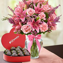 Pink bouquet of roses, lilies and carnations in a glass vase beside a heart box of chocolate strawberries