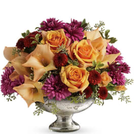 Low centerpiece of orange roses, peach lilies, and purple chrysanthemums in a silver pedestal bowl