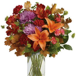 Autumn arrangement of orange lilies, purple mums, and red flowers with brown leaves in a tall glass vase