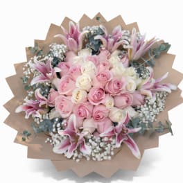 Round bouquet of pink and white roses with pink lilies and baby's breath wrapped in tan paper.