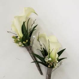 Two white calla lily boutonnieres with dark green leaves and ribbon-wrapped stems