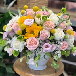 Rounded arrangement of pink and white roses with yellow sunflowers in a white ceramic vase