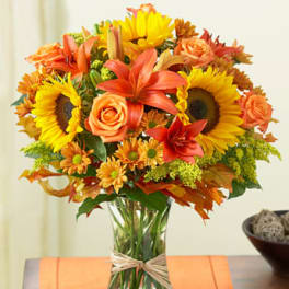 Mixed bouquet of orange lilies, roses, and yellow sunflowers in a clear glass vase