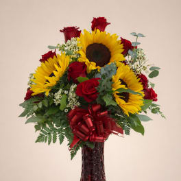 Tall arrangement of yellow sunflowers and red roses in a red glass vase with a red bow.