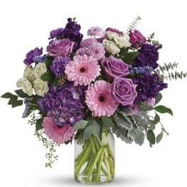 Arrangement of lavender roses, pink gerbera daisies, and purple blooms in a clear glass vase