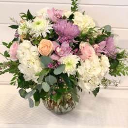 Pastel arrangement of white hydrangeas, pink roses, and lavender mums in a clear glass vase