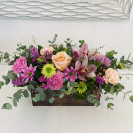 Low arrangement of peach roses, pink carnations, green mums, and orchids in a wooden box