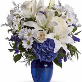White lilies, roses, and daisies with blue hydrangea in a cobalt blue glass vase.