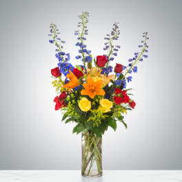 Tall mixed arrangement with orange lilies, red and yellow roses, and blue delphinium in a clear glass vase.