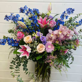 Tall mixed arrangement of pink lilies, roses, orchids and blue delphinium in a clear glass vase