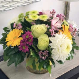 Low round bouquet of white and yellow flowers with green and purple accents in a clear glass vase