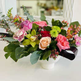 Colorful arrangement of pink and peach roses, carnations, green mums, and an orchid in a white bowl vase.