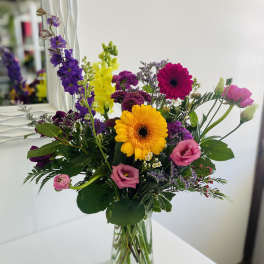 Tall mixed bouquet of yellow and pink gerbera daisies, purple blooms, and pink lisianthus in a clear glass vase.