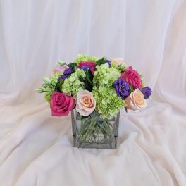 Mixed pink, purple, and green flower arrangement in a square glass vase