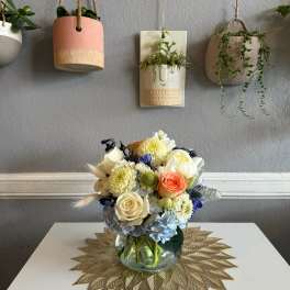 Mixed bouquet in a clear glass vase with cream, peach, blue, and yellow flowers.