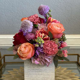 Mixed bouquet of pink, peach, purple, and red flowers in a square vase