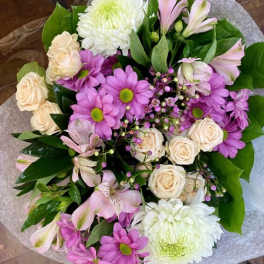 Hand-tied bouquet of cream roses, white mums, and purple daisies wrapped in tissue paper
