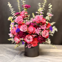Vibrant arrangement of pink roses and red carnations with purple accents in a black cylinder vase