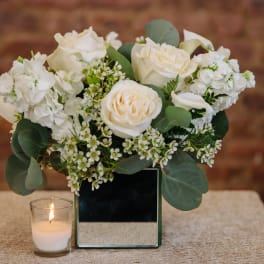 White rose arrangement in a mirrored square vase with a small candle beside it