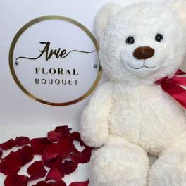 White teddy bear with a red ribbon beside rose petals and a floral bouquet sign