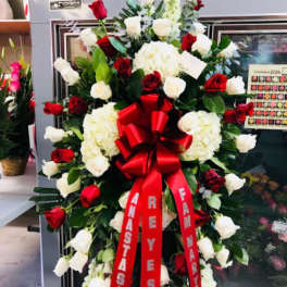 Tall standing spray of red and white roses with white hydrangeas and a large red bow on an easel.