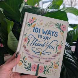 A hand holds a floral gratitude book in front of large green leaves.