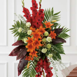 Tall standing spray of red gladiolus, orange lilies, and green carnations on an easel beside a casket.