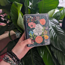 Hand holding a decorative tin box in front of large green leaves