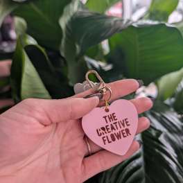 Hand holding a pink heart keychain in front of large green leaves