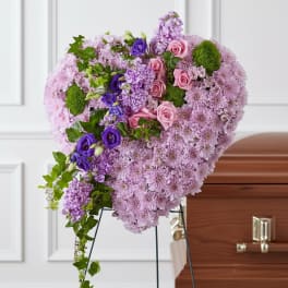 Heart-shaped pink and purple floral funeral spray on a stand beside a casket