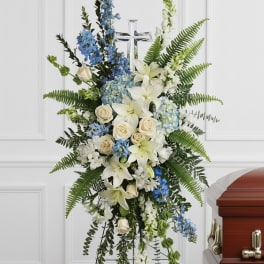 Tall standing spray of white lilies, cream roses, and blue flowers with a cross on an easel