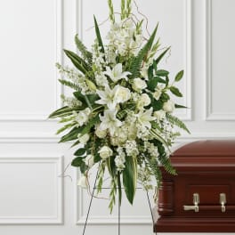 Tall white lily and rose standing spray on an easel beside a wood casket