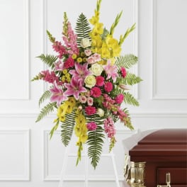 Standing spray of pink lilies, roses, and yellow flowers on a white easel beside a wooden casket.