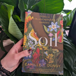 Hand holding a floral-covered book in front of large green leaves.