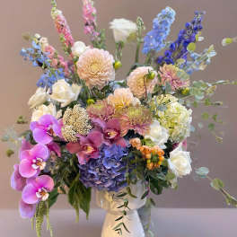 Mixed pastel bouquet in a white vase with orchids, roses, hydrangea, and tall blue blooms