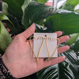 Hand holding gold heart-shaped earrings in front of large green leaves.