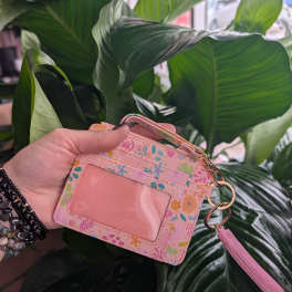 Hand holding a pink floral ID wallet in front of large green leaves.