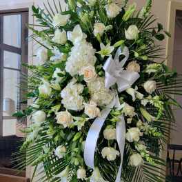 Tall standing spray of white roses, lilies, hydrangeas, and palm fronds with a white bow on an easel.