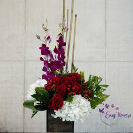 Tall floral arrangement with red roses, white hydrangeas, and purple orchids in a dark vase