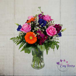 Mixed bouquet of pink roses, orange gerbera daisies, and blue blooms in a glass vase