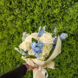 Hand-tied bouquet of white roses and blue flowers in cream wrap with ribbon, held against a green wall.