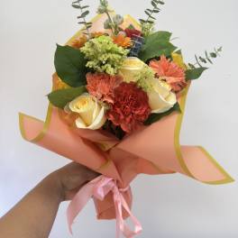 Handheld bouquet of roses, carnations, and orange flowers wrapped in peach paper