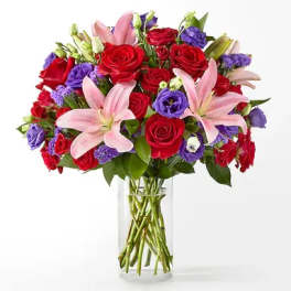 Vase arrangement of pink lilies, red roses, and purple blooms in a clear glass cylinder
