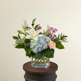 Low mixed arrangement with white lily, blue hydrangea, peach carnations, and purple flowers in a glass vase