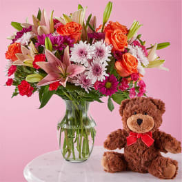 Mixed bouquet in a glass vase beside a teddy bear with a red bow